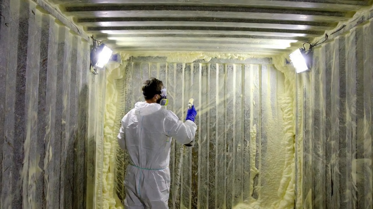 Worker in protective suit applying closed-cell spray foam insulation inside a container